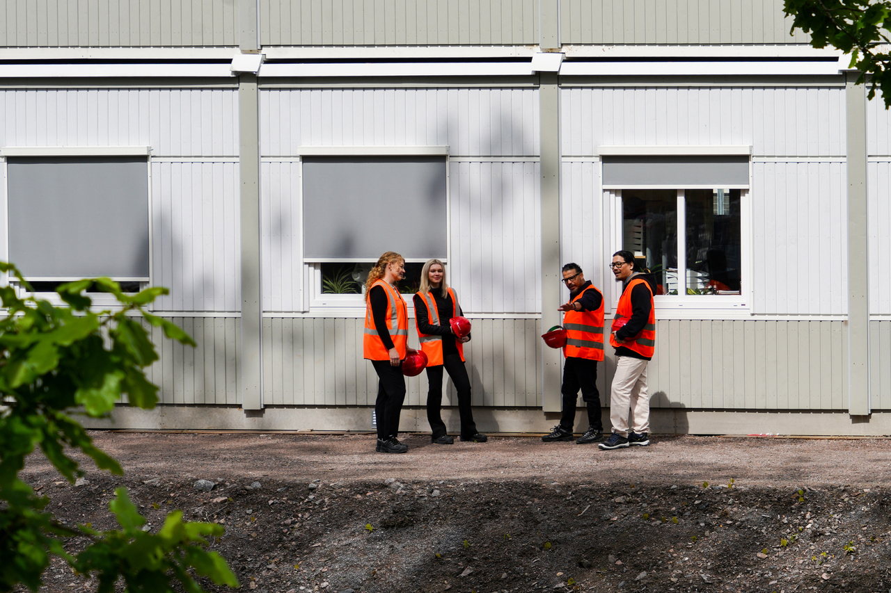 Fyra personer i varselvästar står utanför kontorsmoduler från Expandia vid Skanskas projektkontor i Karlstad. De håller röda skyddshjälmar och samtalar framför modulfasaden med fönster, i en miljö som visar övergången mellan kontor och byggarbetsplats.