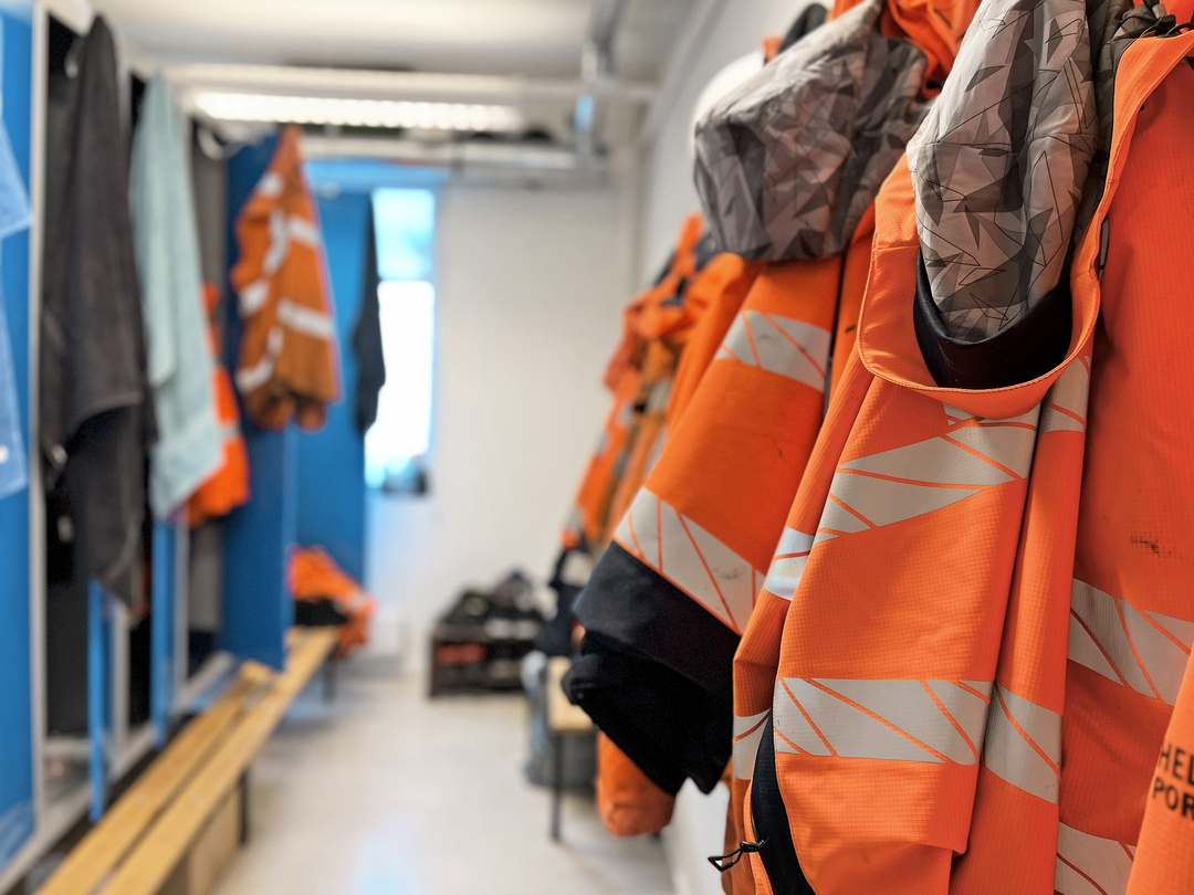 Omklädningsrum med blå skåp och bänkar, där orange varseljackor med reflexband hänger längs väggen; skor och arbetskläder syns i bakgrunden i ett ljust och funktionellt utrymme.