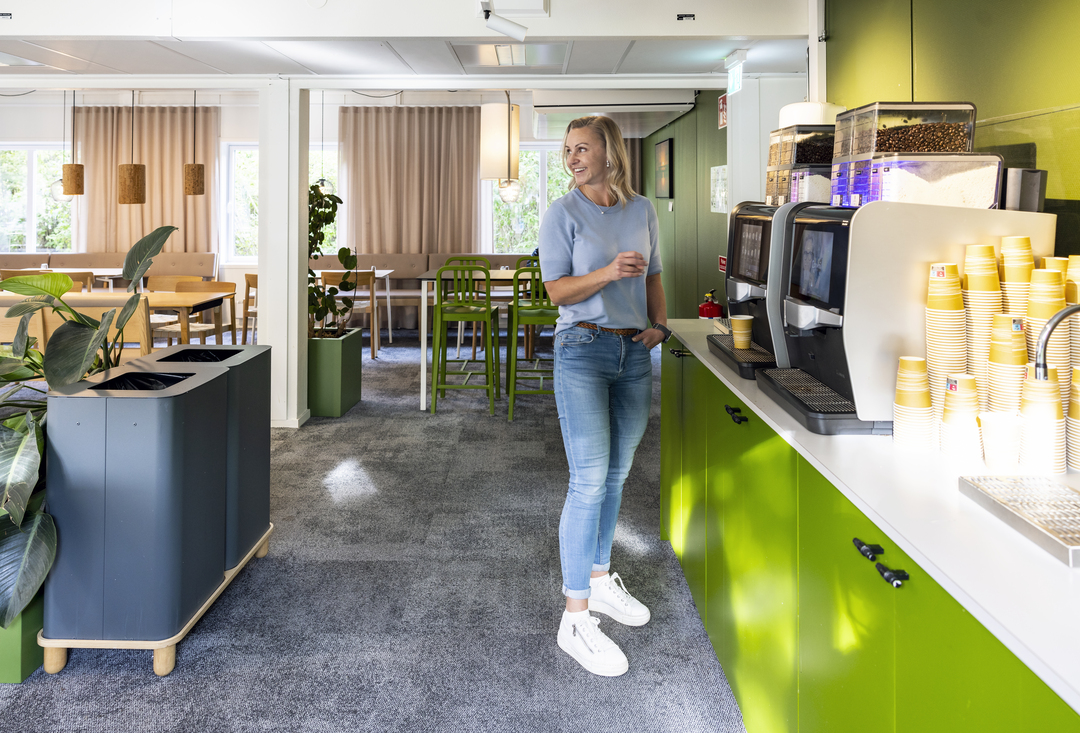 Pausyta och kaffestation i kontorsmoduler hos Polypeptide med gröna skåp, kaffemaskiner, sittgrupper, växter och mjuk belysning där en medarbetare tar kaffe i en trivsam arbetsmiljö – kontorsmoduler från Expandia.