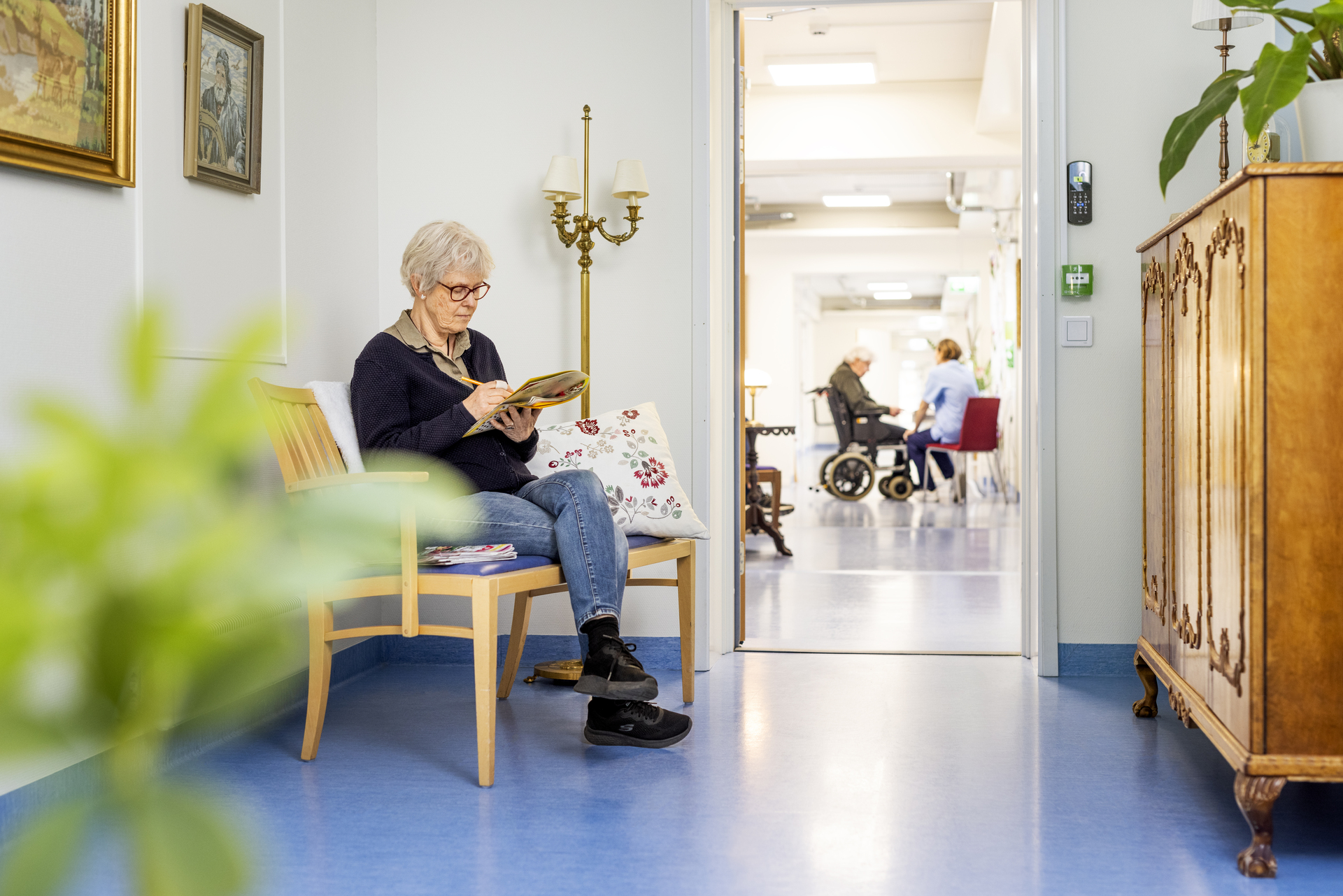 En boende sitter i en korridor och läser en tidning vid Borggården demensvård i Olofström. I bakgrunden syns en annan boende och omsorgspersonal i samtal, i en ljus miljö som ingår i en modullösning från Expandia.